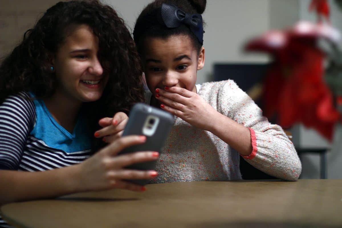 Two young girls laugh while looking at a smartphone, one pointing at the app and the other covering her mouth in surprise.