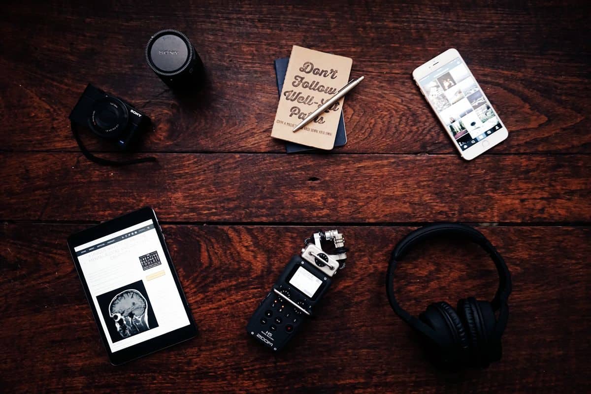 A wooden desk displays multiple tools for building mobile apps, including a tablet, a handheld voice recorder, a smartphone, a camera, a pair of headphones and a small notebook.