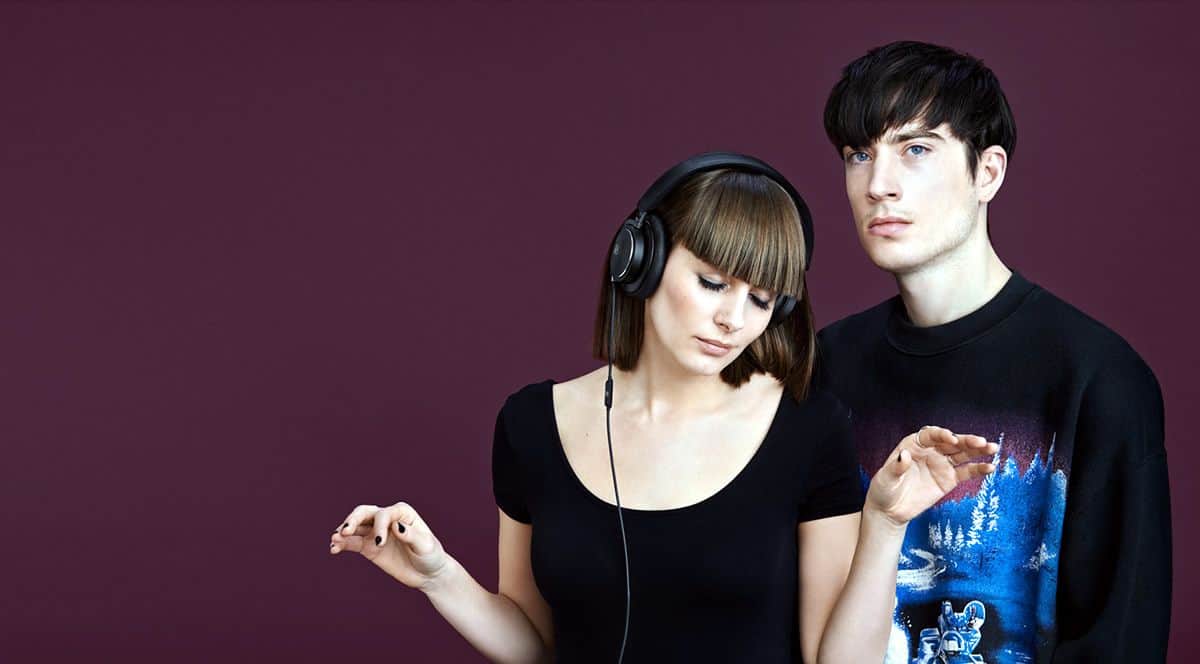 Young woman listening to music with the BeoPlay H6 in black and a young man standing next to her facing the camera