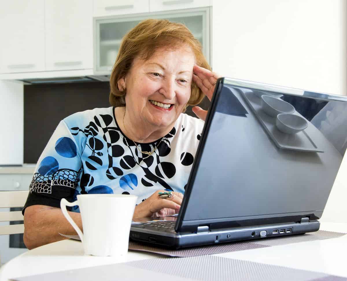 A senior woman sits in front of her laptop smiling.
