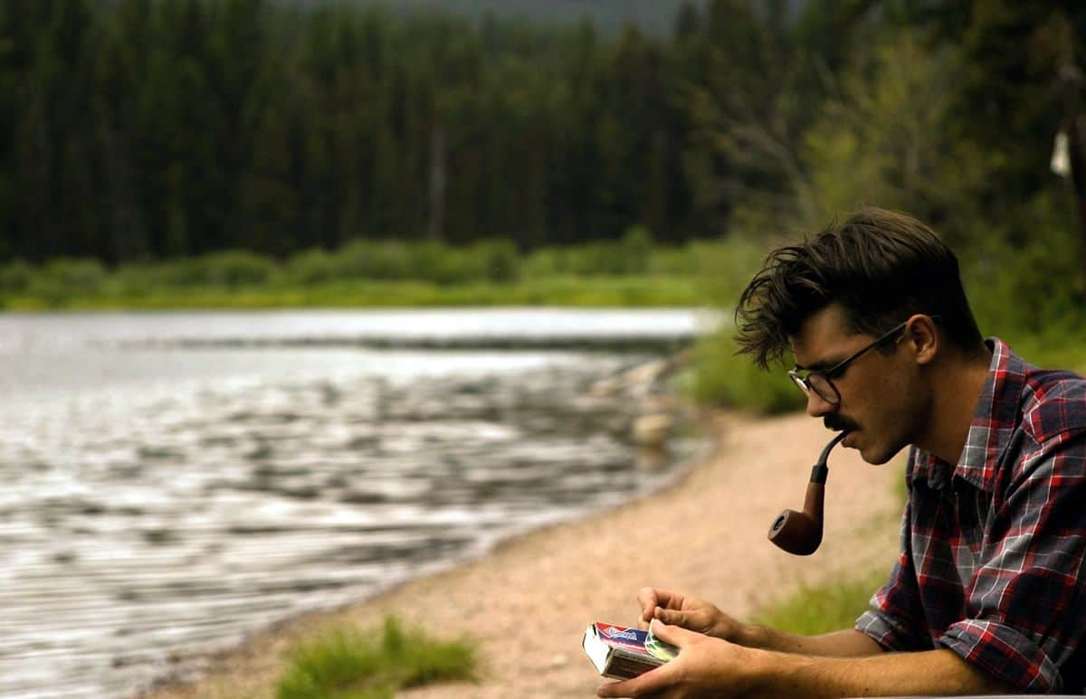 A young bespectacled man with a pipe and a book sitting by the woods and the lake, representing the idea of the modern day guerrilla UX designer