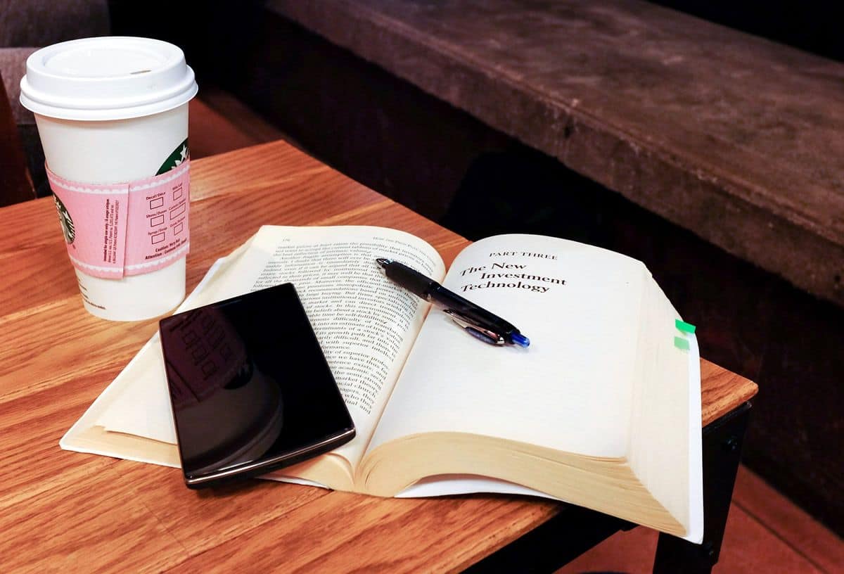 A mobile phone sitting on top of a heavy book next to a Starbucks coffee cup.