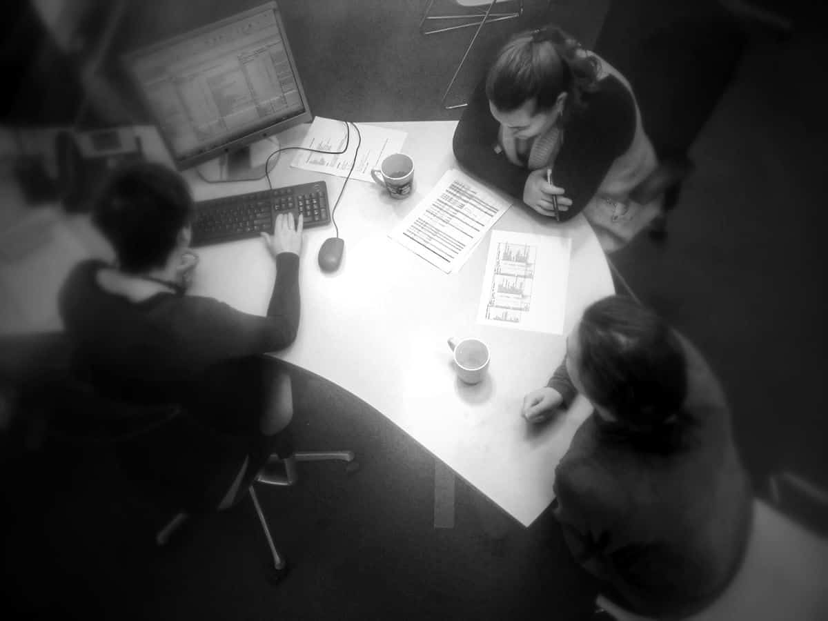 A top-down view of a mobile UI design team of three people sitting at a desk with cups of coffee, notes and a desktop computer, discussing budget and scope of a project.