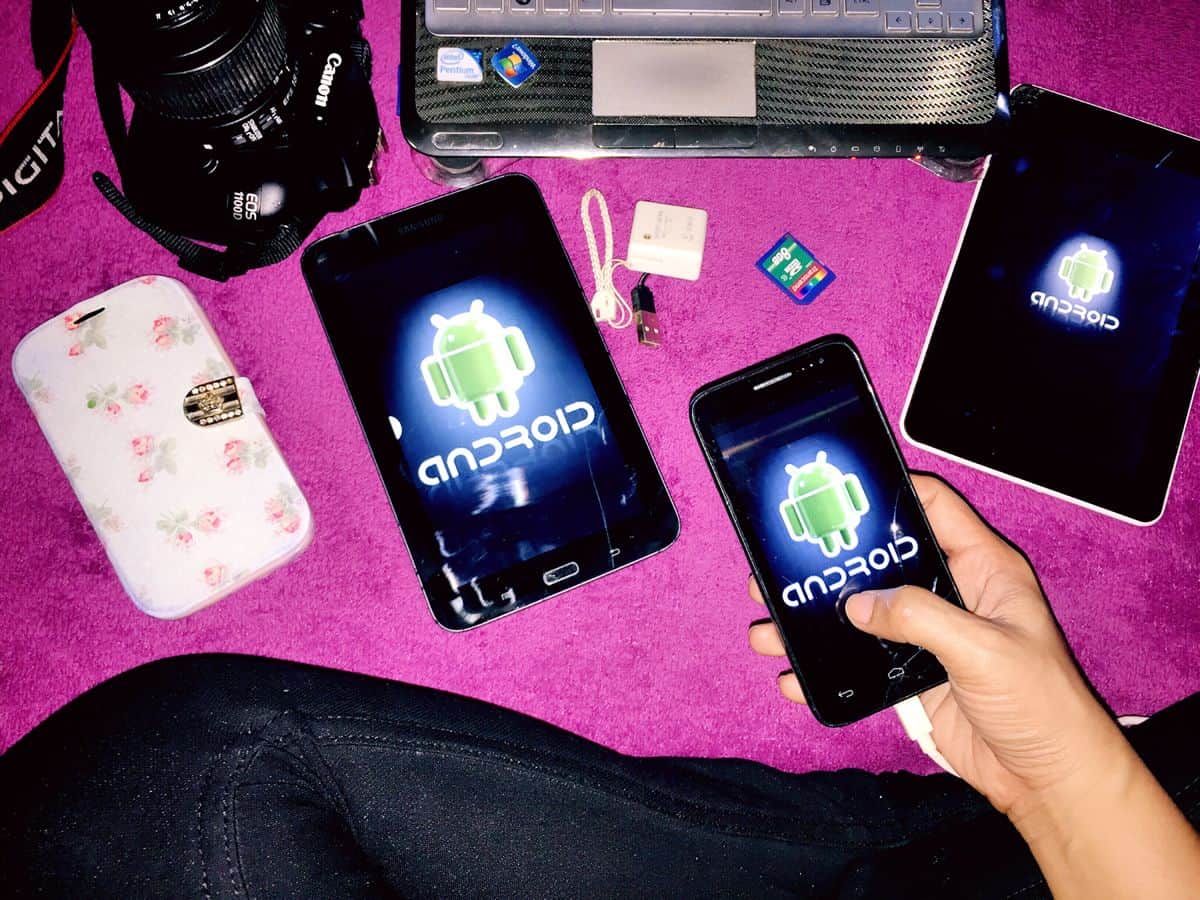 An Android UI designer sits cross-legged on the floor holding an Android smartphone. On the floor are two Android tablets of different sizes and a larger Android smartphone, in addition to a camera and laptop.