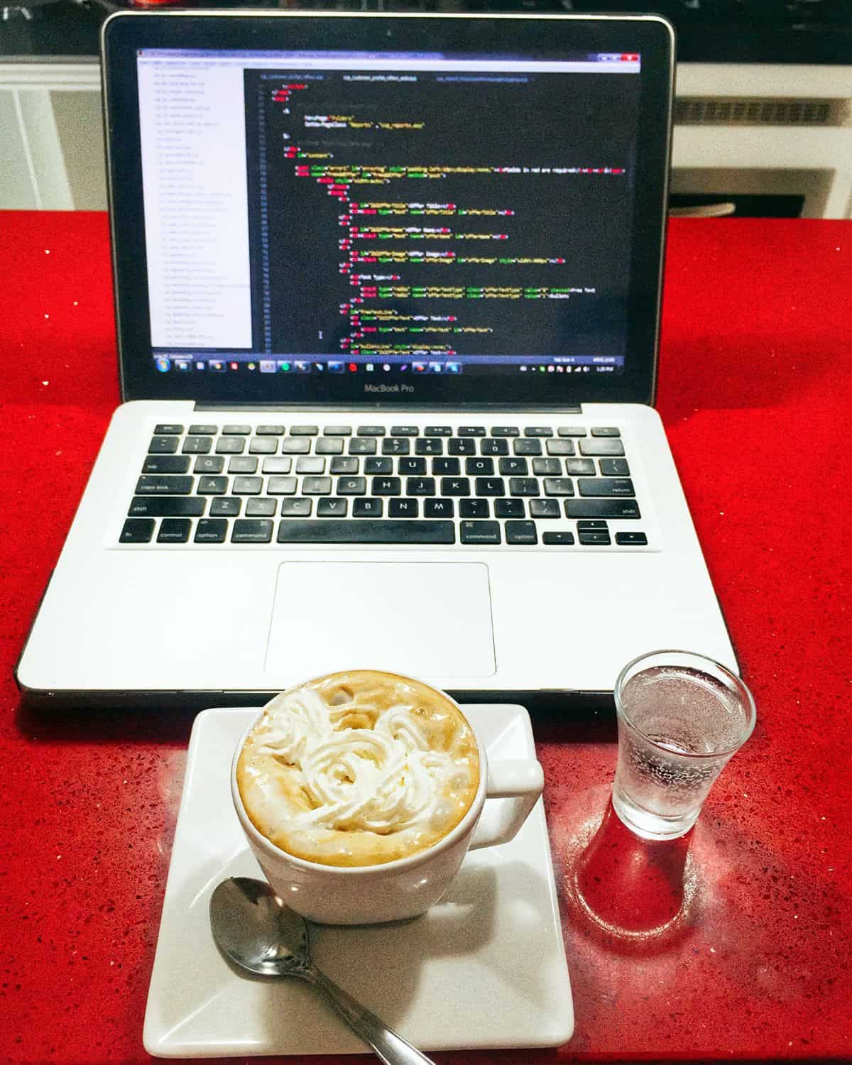 A cappuccino and a small glass of water sit in front of a developer’s laptop, where a code editor is open.