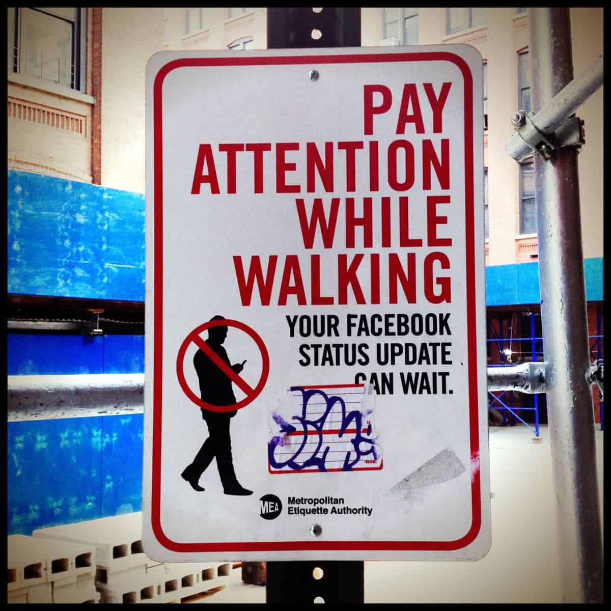 A street sign shows the silhouette of a person walking while using his mobile phone, with the message, “Pay attention while walking. Your Facebook status update can wait.”