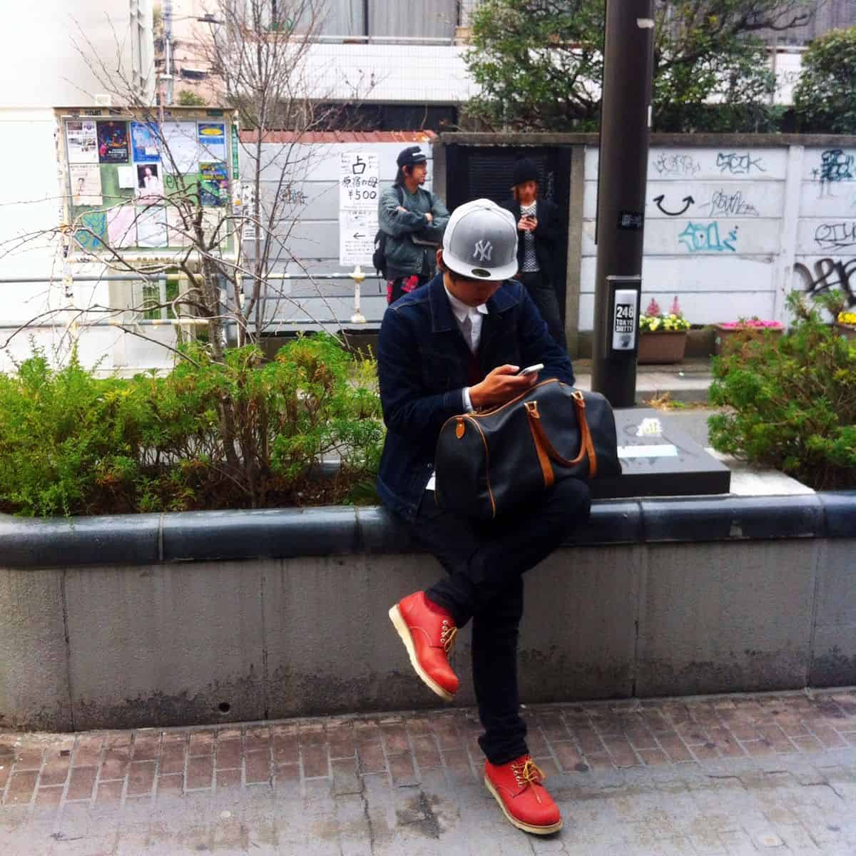 A man in trendy business attire and a New York Yankees hat sits at a bus stop, using his smartphone while waiting for transportation.