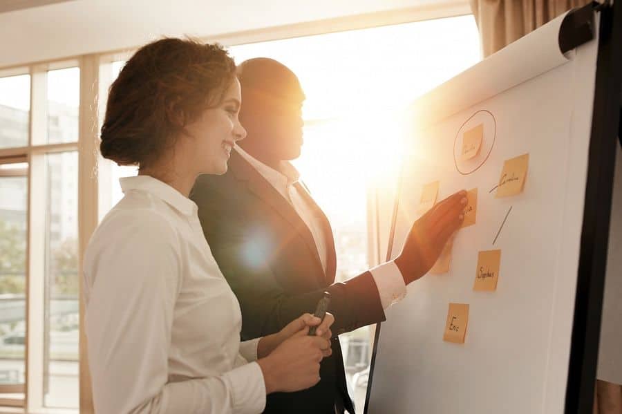 Two coworkers stand in front of a white flipboard, adjusting sticky notes to edit a user onboarding flow.