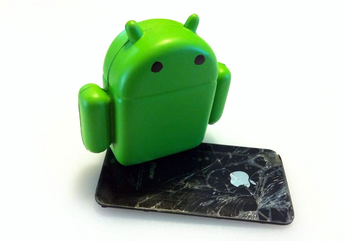 A figurine of the Android mascot stands on top of an iPhone with shattered glass.