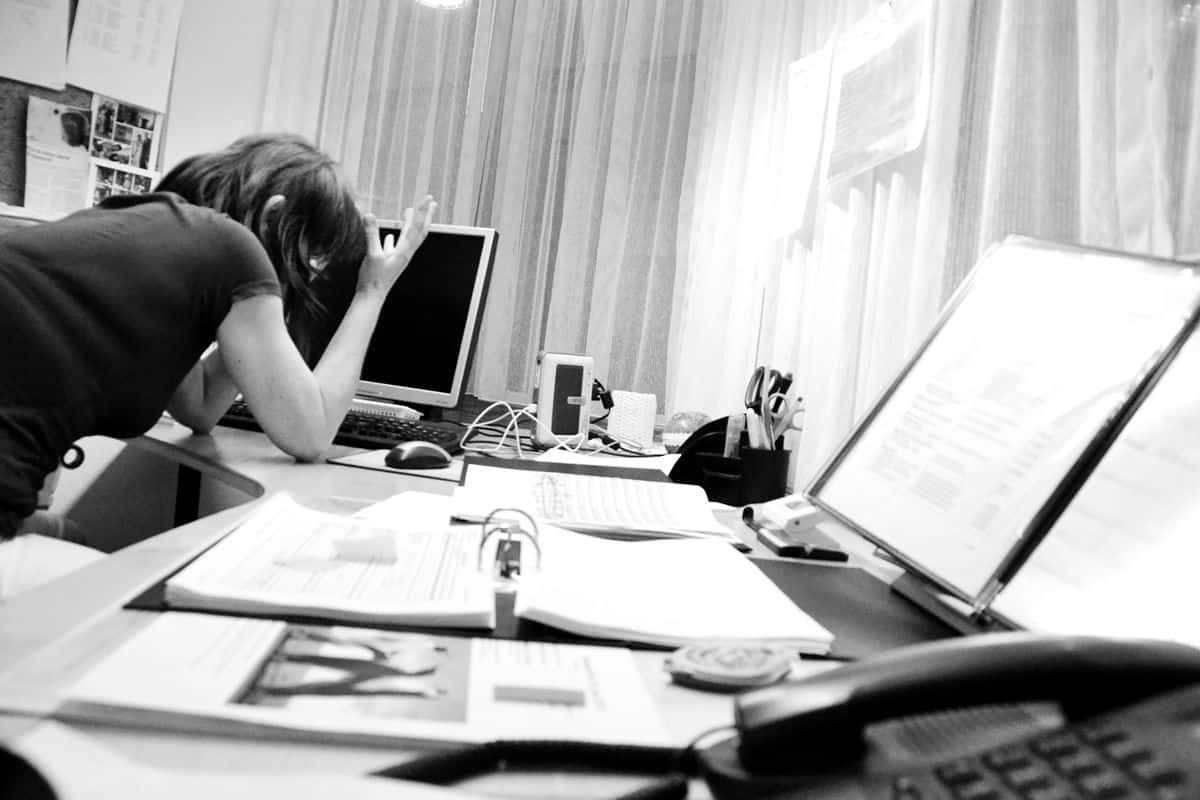 A frazzled office worker, with multiple binders and notebooks open on her desk, props her elbows on the desk and gestures in frustration.