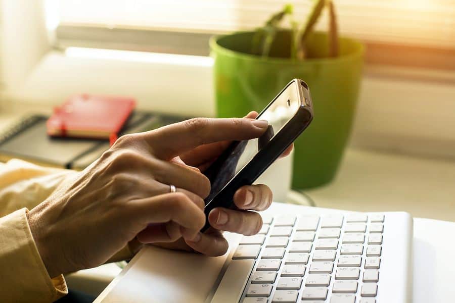 A closeup of a woman’s hand interacting with a mobile phone, resting just in front of a desktop computer keyboard.