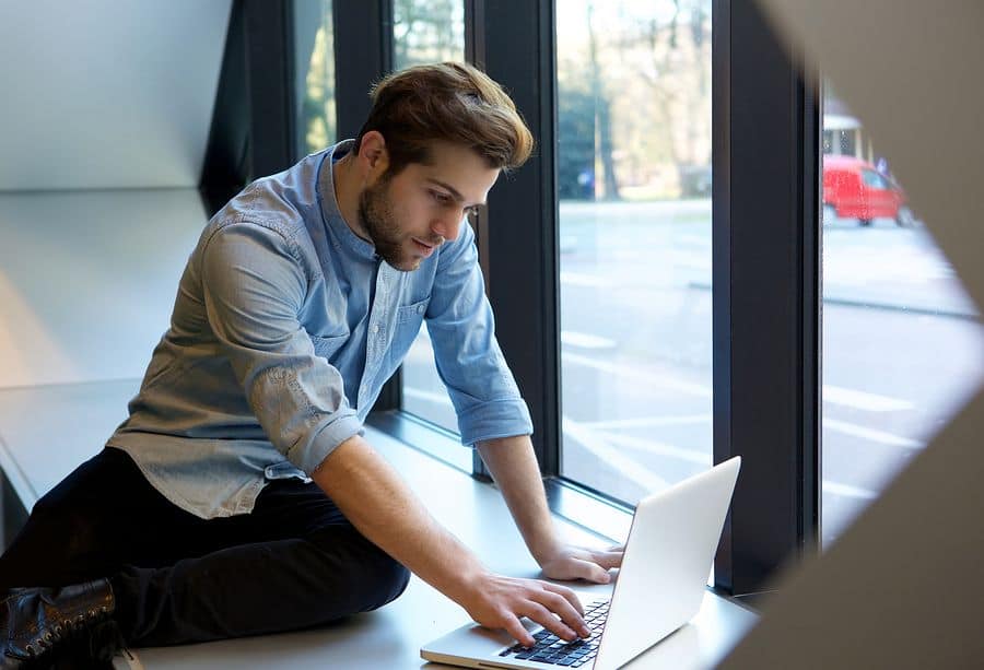 A man in business attire works remotely from his laptop while sitting on a large window sill.