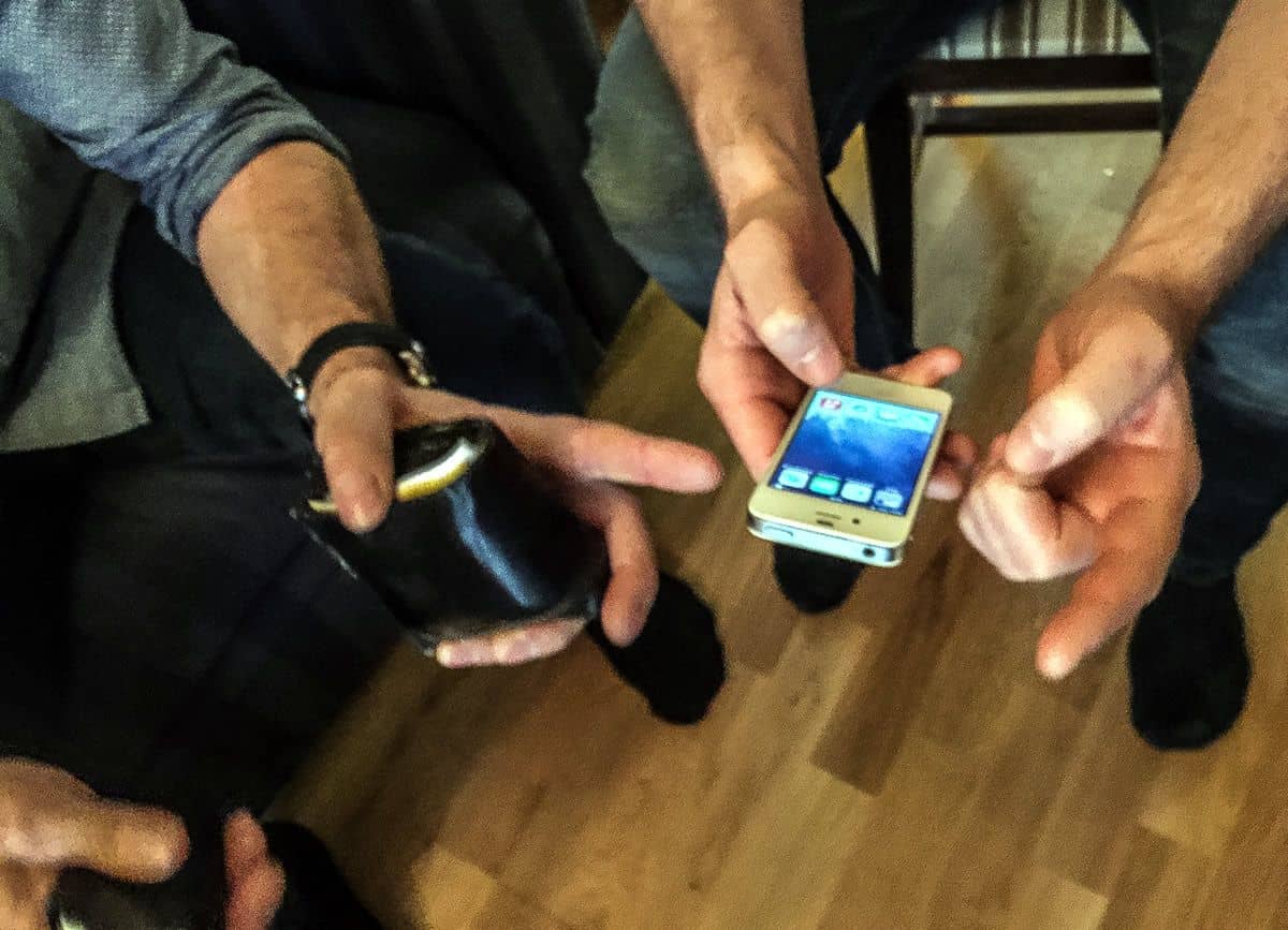 A close up on two men’s hands as they hold a conversation. One man is holding a smartphone and pointing while the other is holding a wallet and pointing at the smartphone.