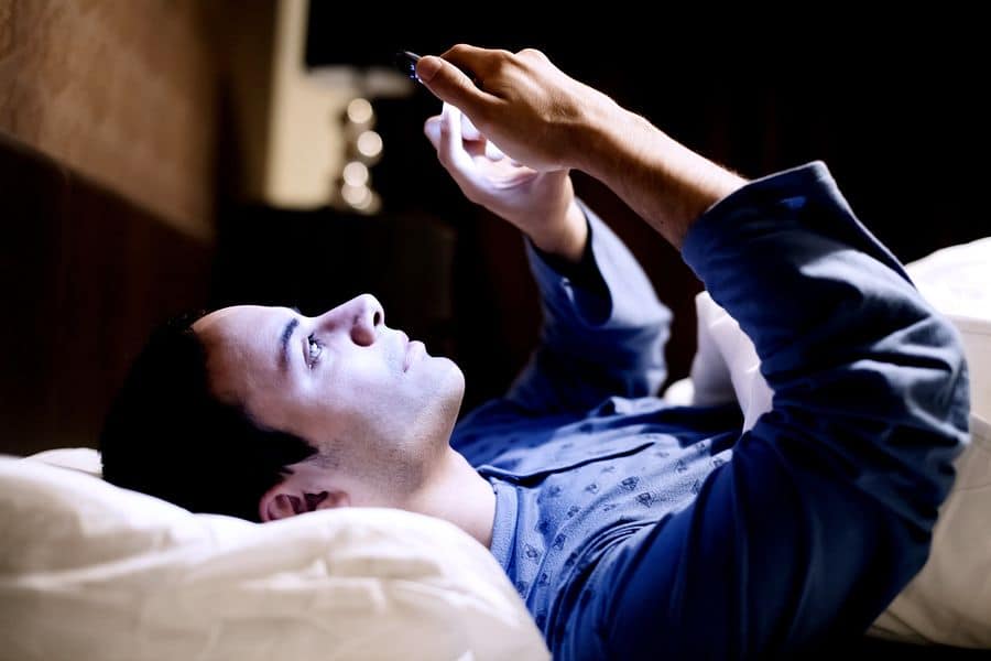 A photo of a man lying in bed in a darkened room, playing a game on his mobile phone.