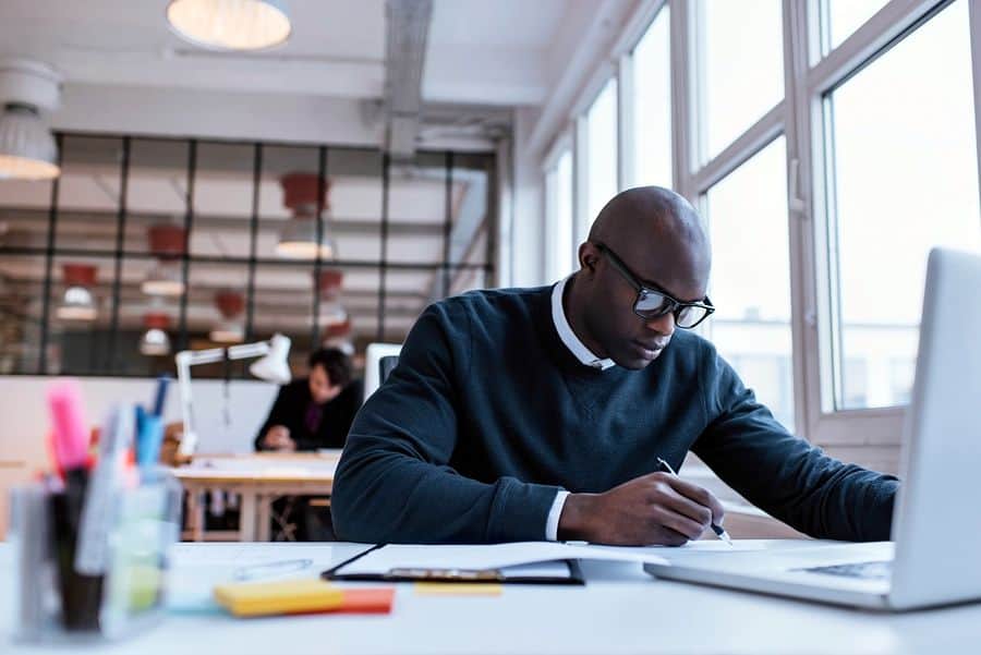 A web app UI designer in a modern agency sits at a white desk in front of his Macbook, sketching out a design using a pen.