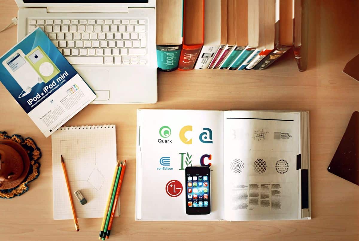 The desk of a mobile and web app UI design student, complete with coding and design textbooks, a graph paper notebook, colored pencils, and MacBook.