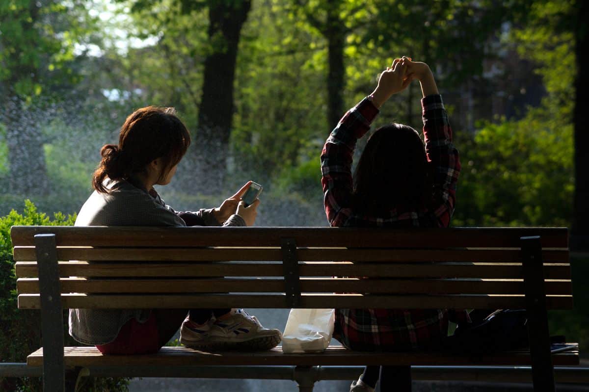 Two women sitting on a park bench, the one on the left using her mobile phone and the other stretching her arms overhead.