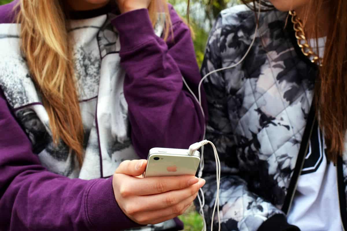 Two young women sitting next to each other sharing a pair of earphones connected to an iPhone.