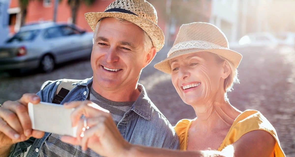 A smiling elderly couple on vacation holding up an iPhone to take a selfie.