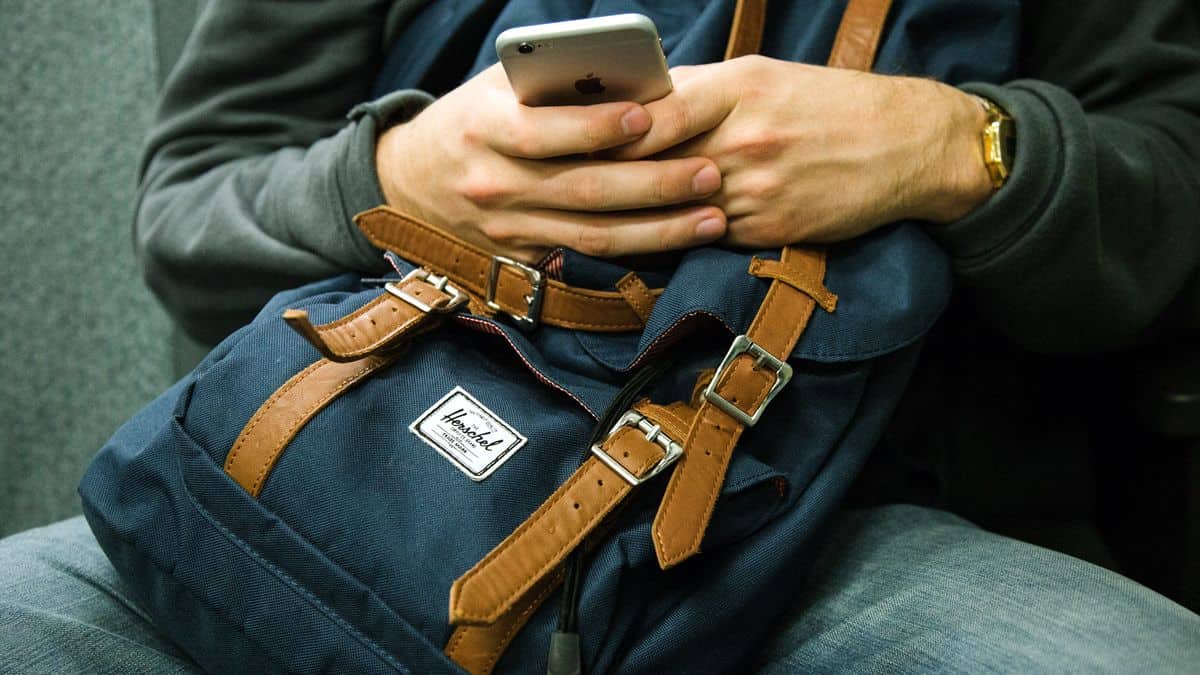 Close up of a seated man using an iPhone with a knapsack clutched to the front. User interviews help designers to understand the user in context.