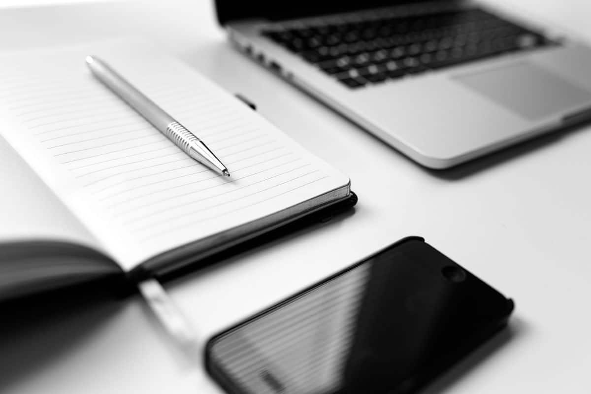 A black and white photo of a pen sitting in an open notebook on a desk with an iPhone and MacBook.