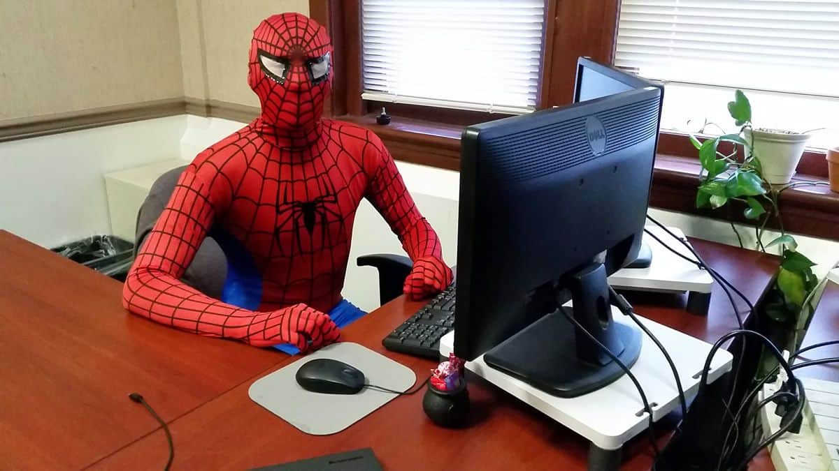 A person in a Spiderman Costume sits behind a computer in an office setting.