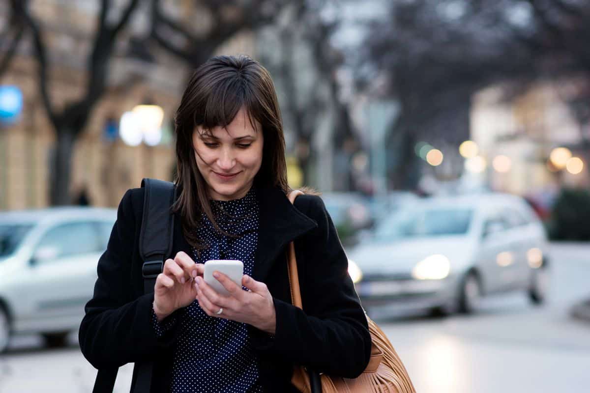 A smiling professional woman walking down a busy street interacts with an app on her phone.