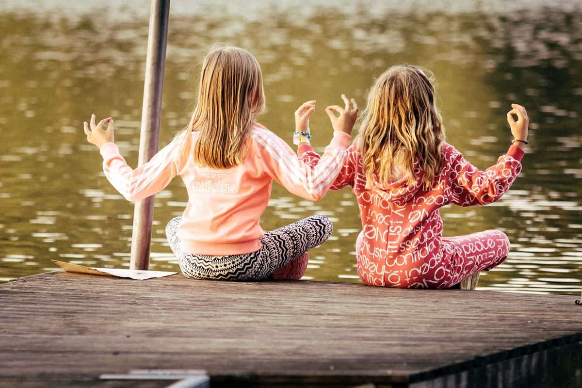 Two young girls sit cross-legged at the edge of a dock in front of a lake, practicing meditation.