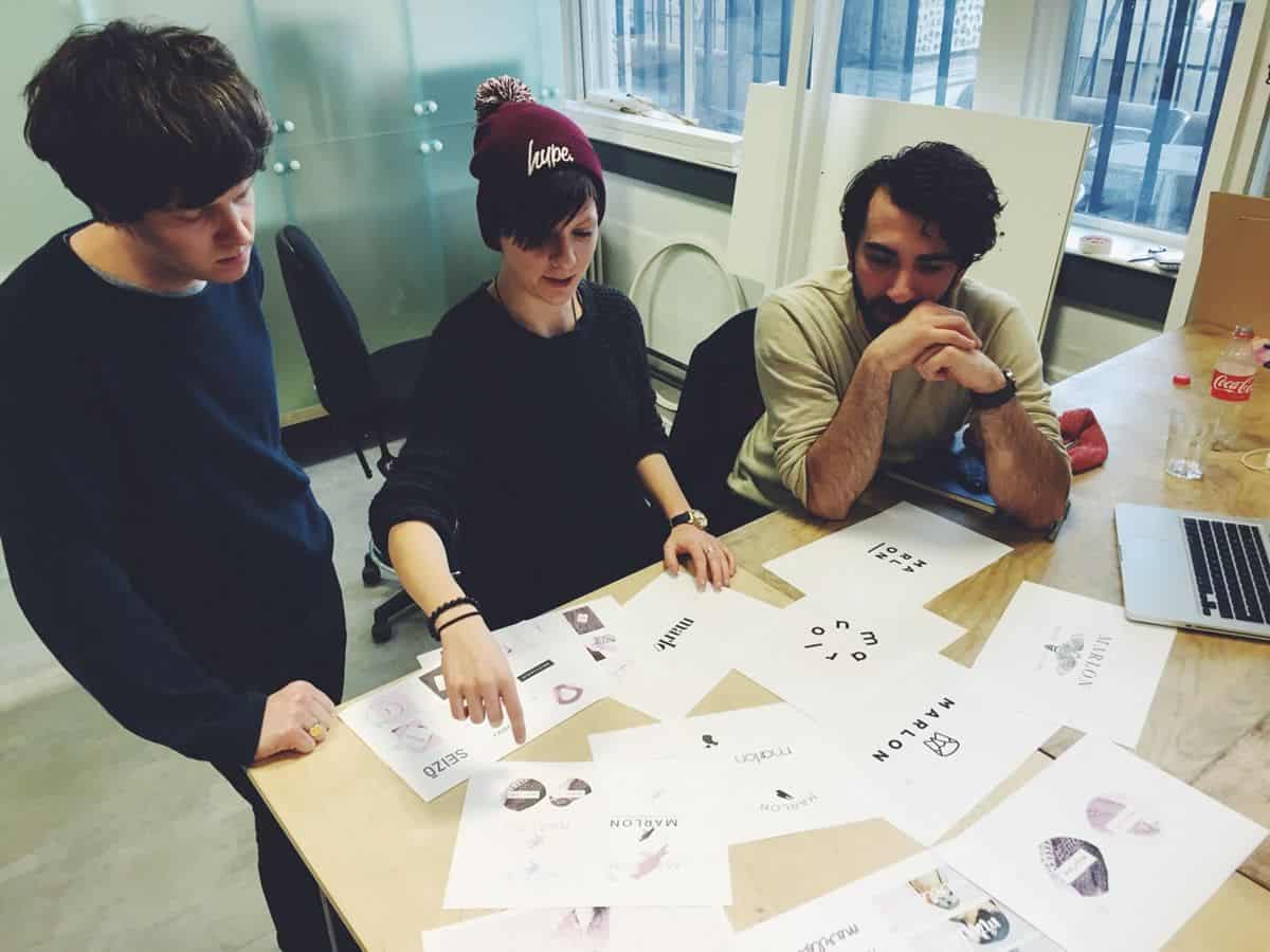 A product design team of three designers at Morrama sits at a desk covered in printed out branding options, discussing their ideas.