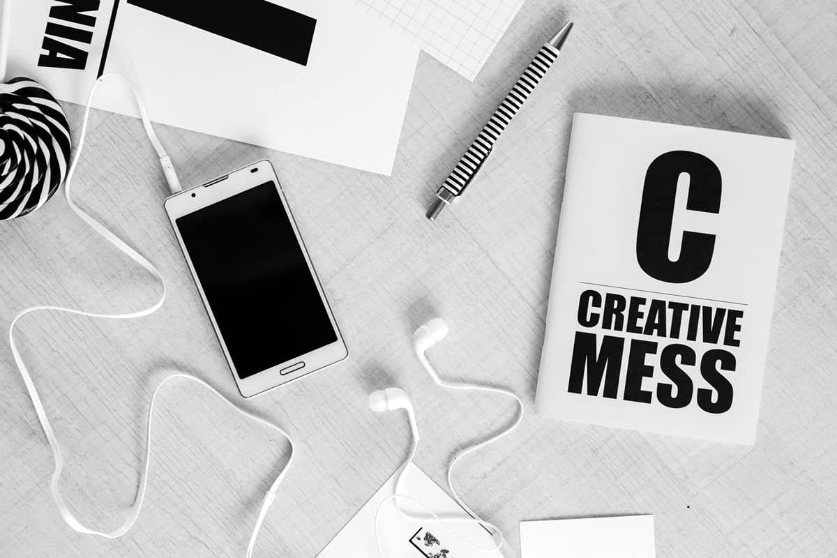 White earphones plugged into a white mobile device on a white table that holds a black and white striped pen and a white booklet with black text that says, “Creative Mess”.