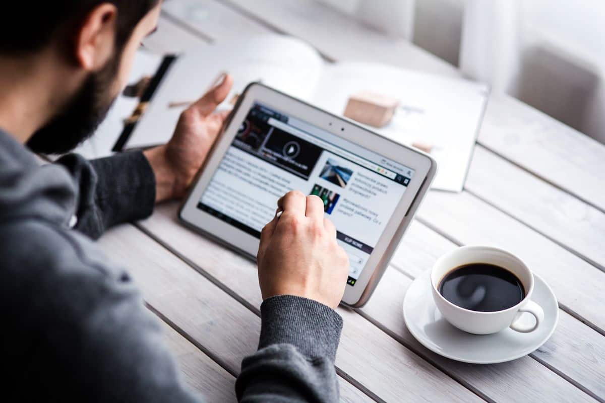 A young man sitting on a wooden bench table using a tablet with a stylus while having a cup of coffee.