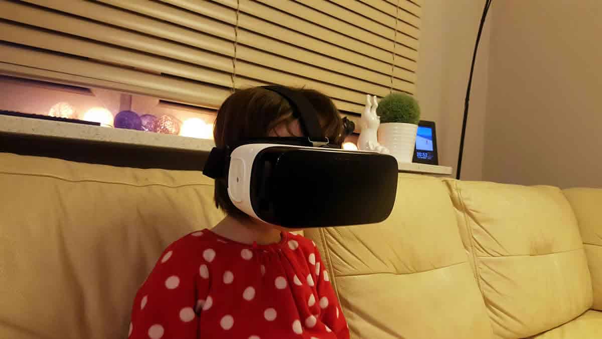 A young girl sits on a couch wearing a Samsung Gear VR virtual reality headset.