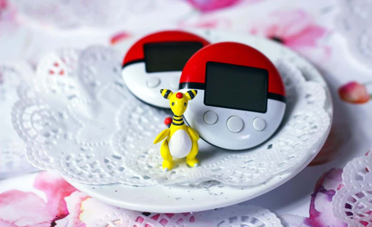 A small Pokemon figurine stands on a small tea dish on top of delicate doilies, in front of two PokeWalkers.