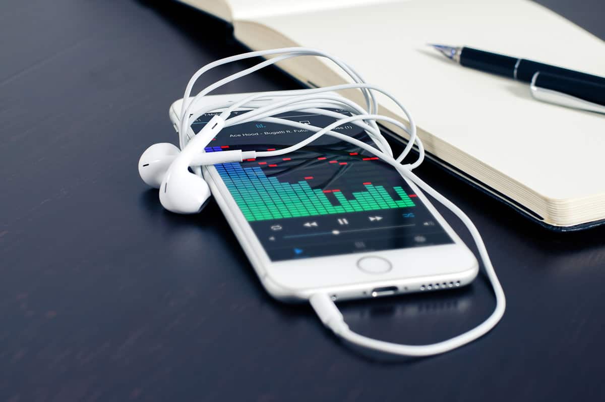 An iphone with earbuds wrapped around it with a music player app on the screen, next to a notebook opened to an empty sheet and a pen.