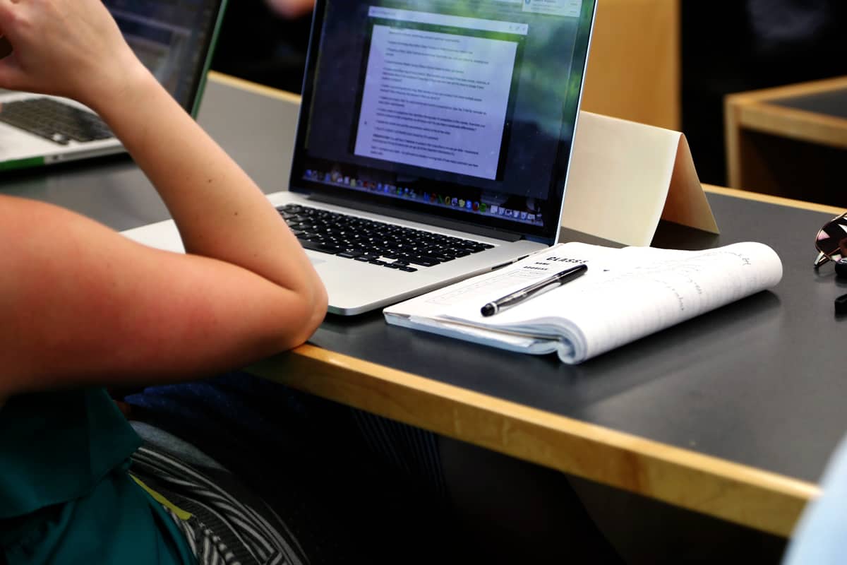 A freelance UX designer reading a document on her MacBook and taking notes in a notebook.