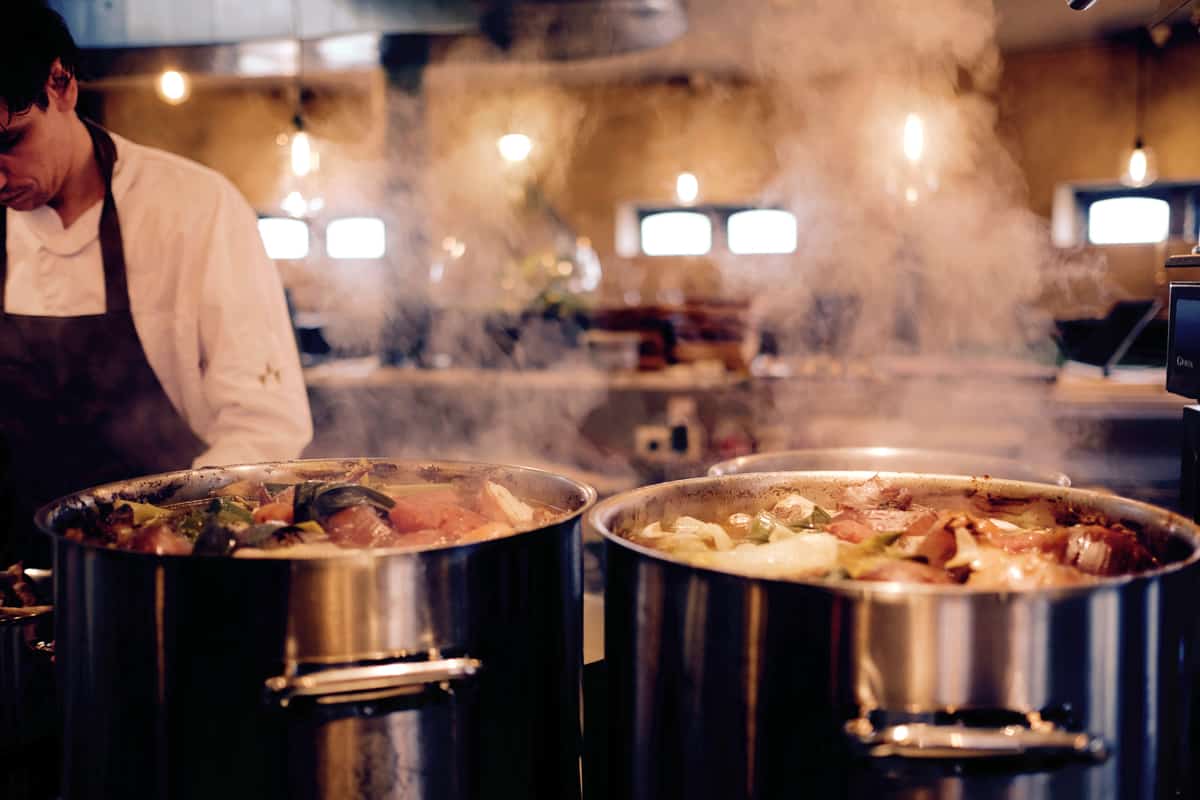 A close-up shot of two huge pots of soup on a stovetop steaming in a kitchen.