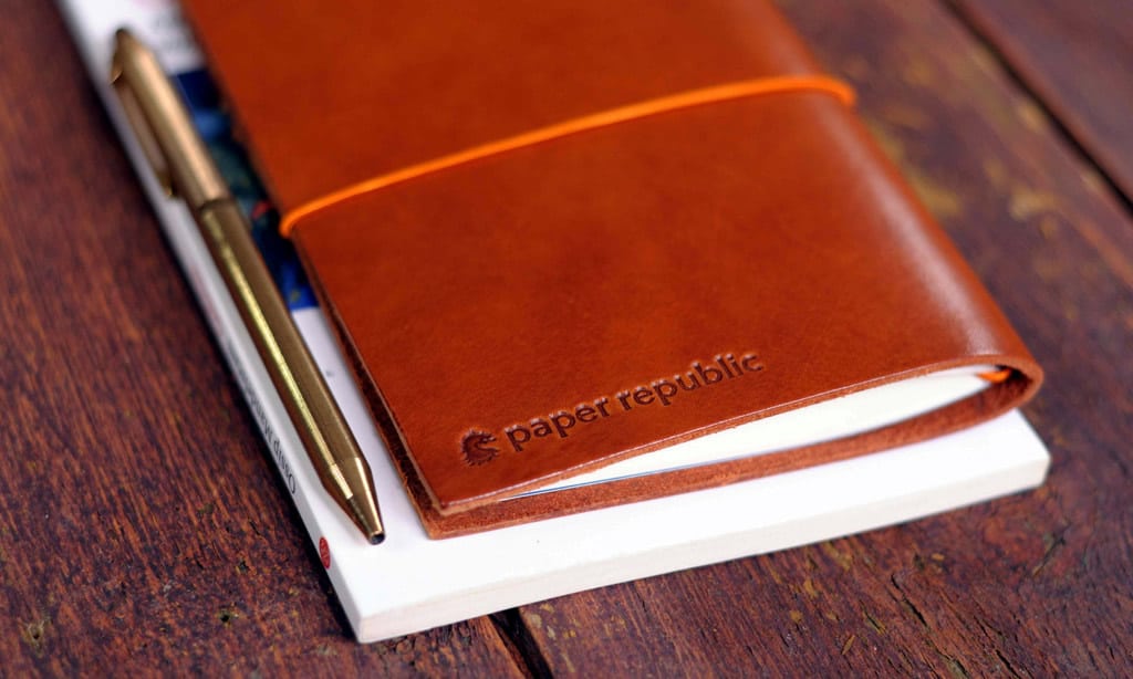 Brown leather cover bearing the name Paper Republic, holding a notebook within and a golden pen.