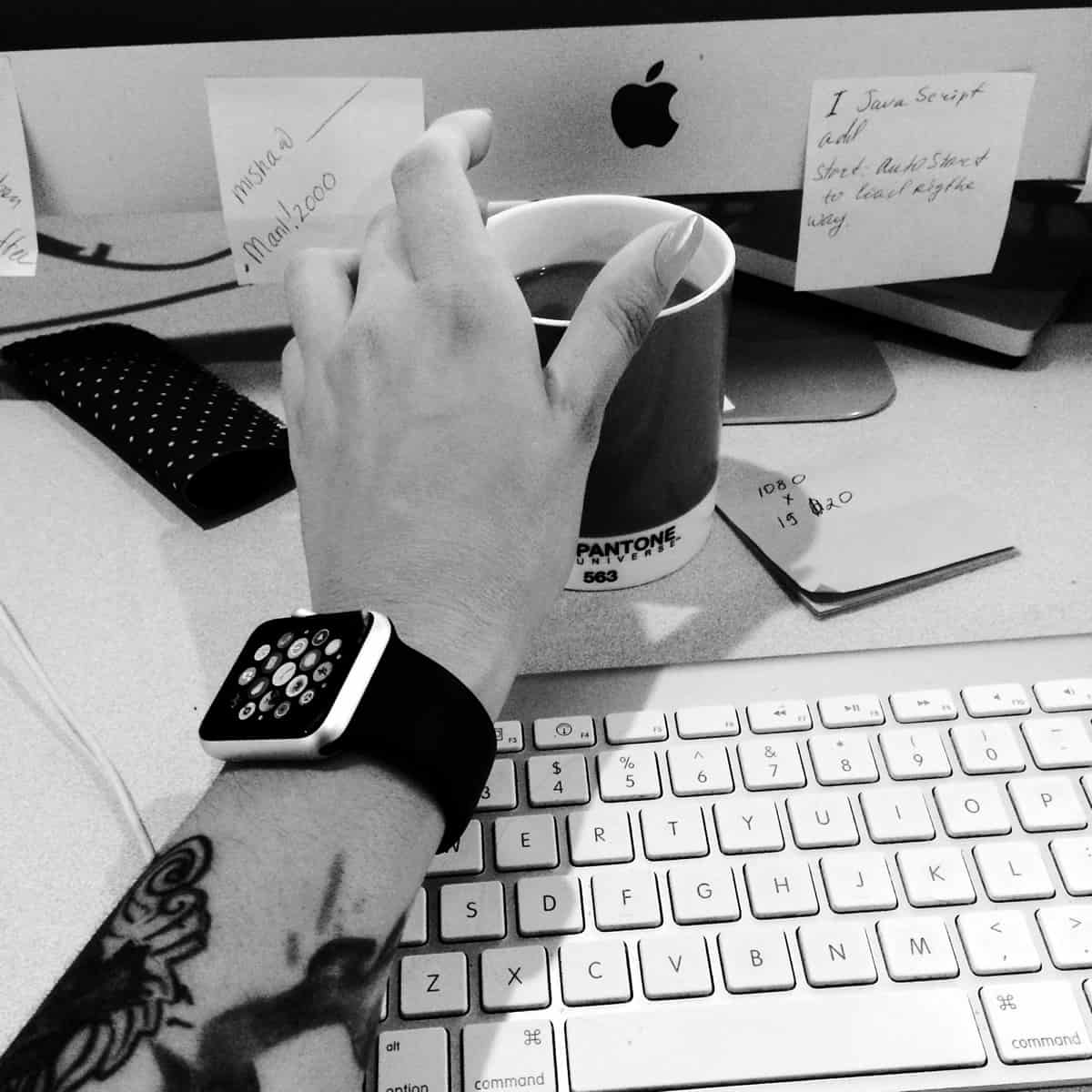 A black and white photo of a mobile app designer reaching for a coffee cup near an iMac monitor while wearing an Apple Watch.