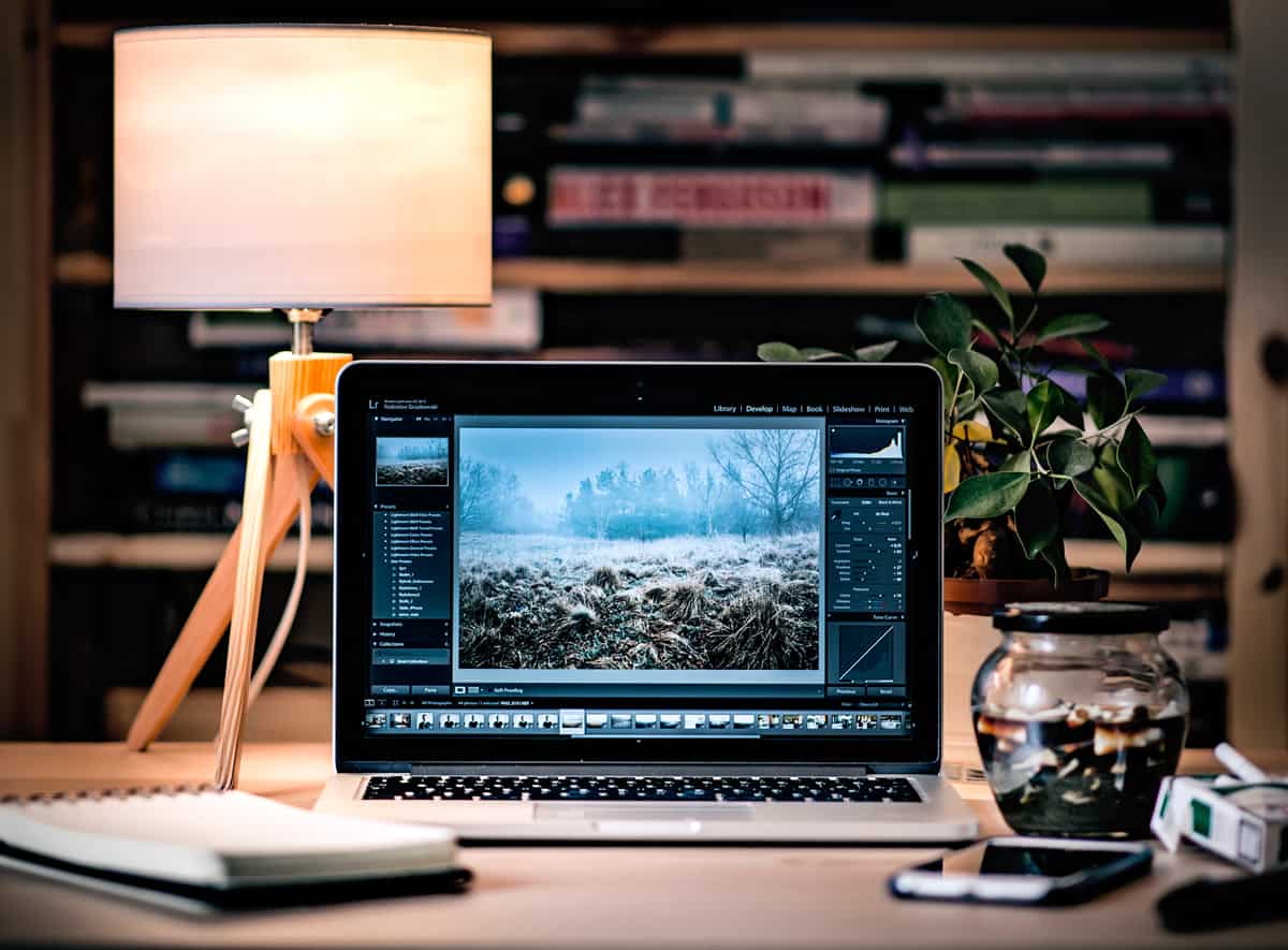 A MacBook opened to a photo editing software on a work desk with a book shelf in the background.