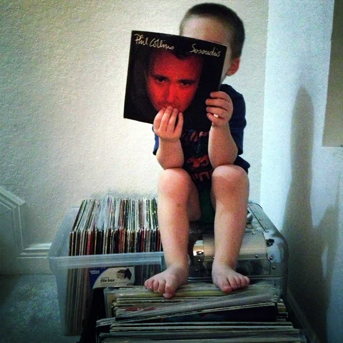 A young boy sits on top of a stack of vinyl, holding up a Phil Collins album.