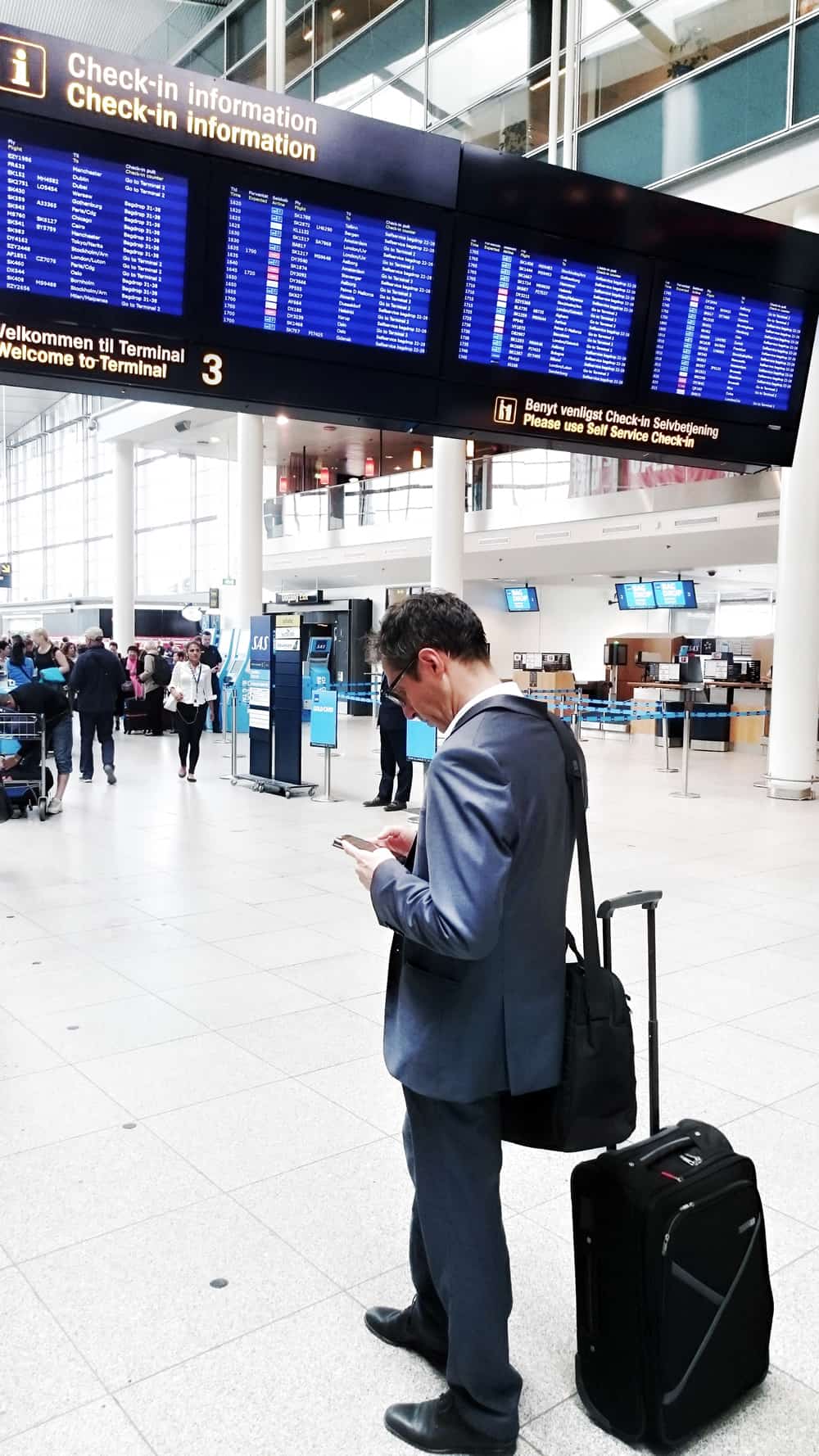 A photo of a man in a suit consulting his smartphone in front of a board detailing arrivals and departures at the airport.
