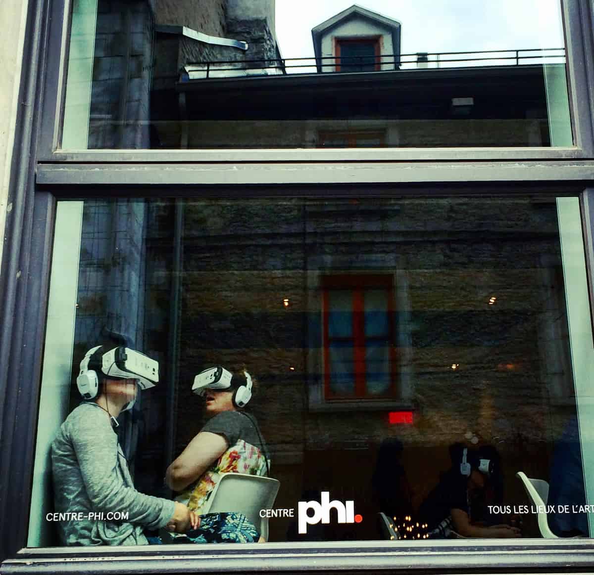 A photo of a couple sitting in a storefront window, each wearing head-mounted virtual reality goggles.