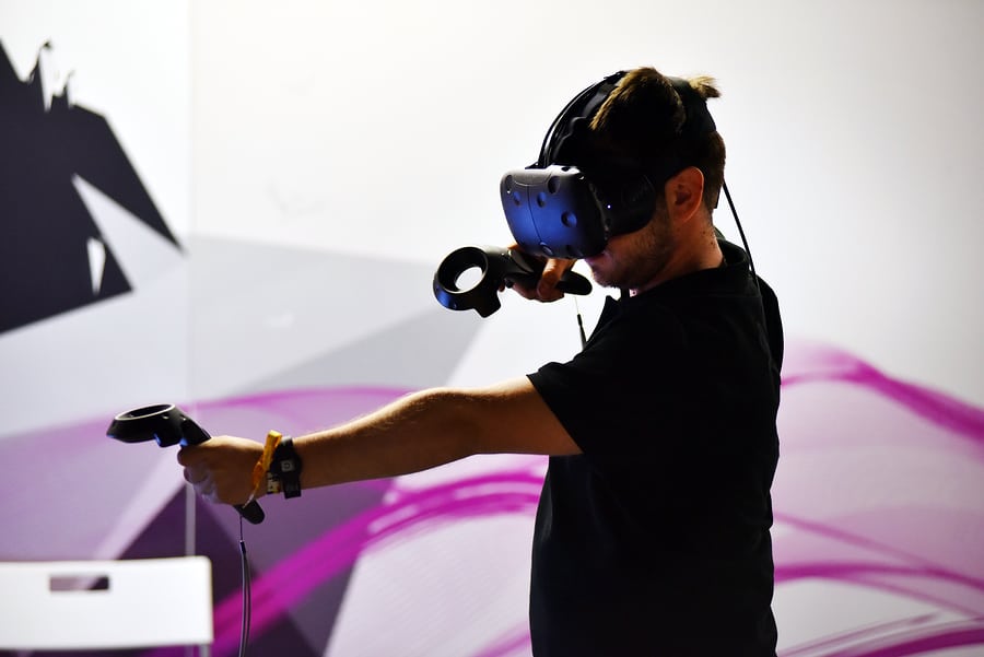 A photo of a man wearing an HTC Vive HMD uses handheld controllers to play a game.