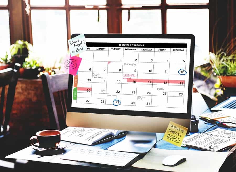 A photo of a desk full of papers and a computer displaying a calendar with Post-Its on its edges. We have some cool apps to help with this.