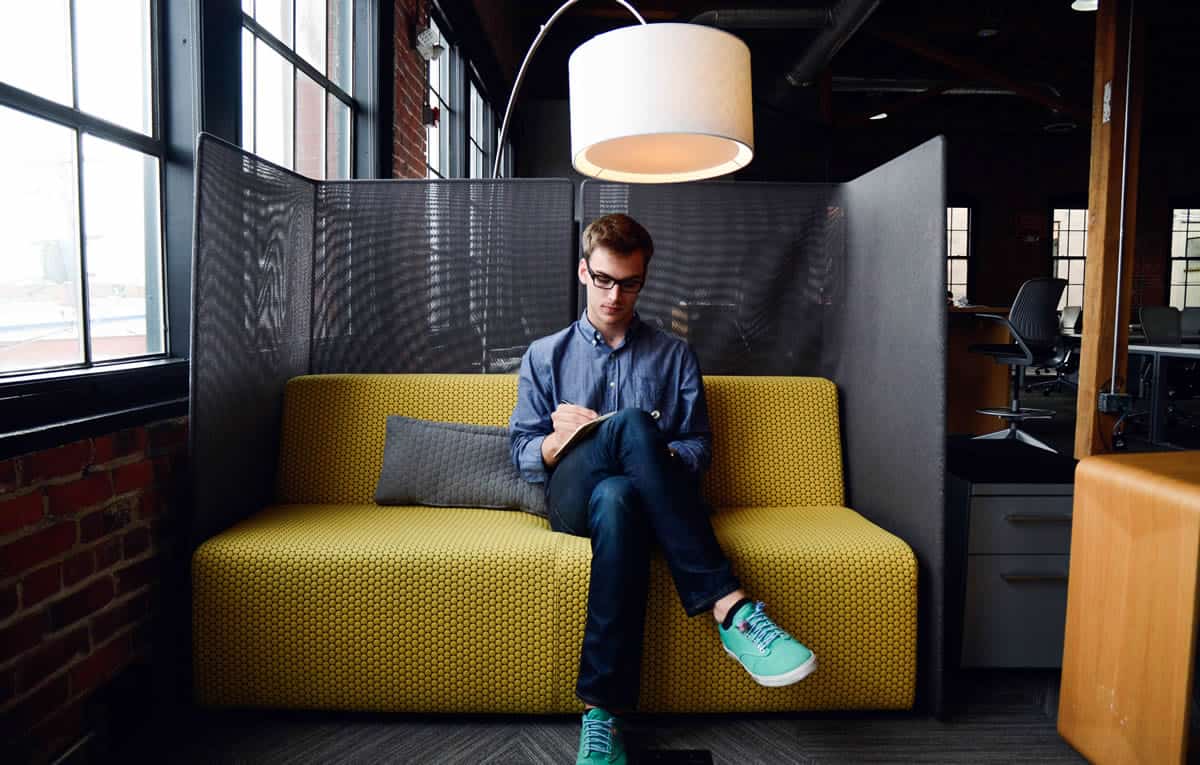 Man writing in a notebook on a couch in the nook of an office.