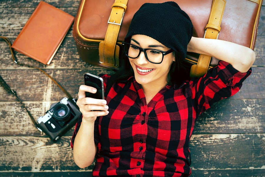 A photo of a woman, smiling and resting her head on a suitcase while looking at her iPhone.