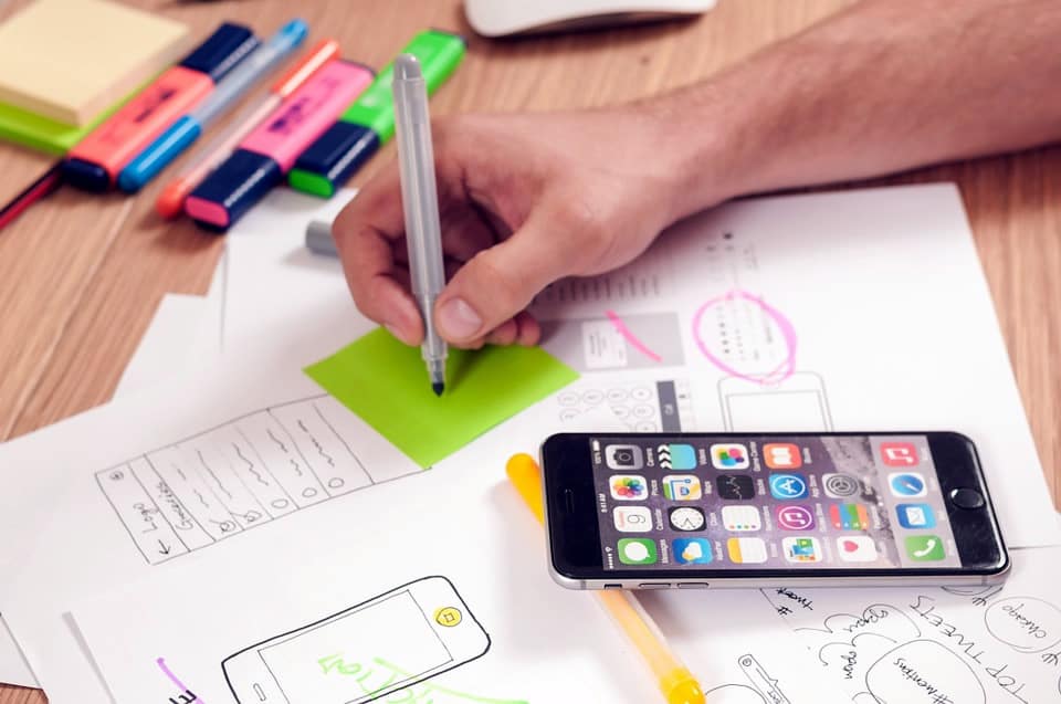 Image of a table, mobile phone and person sketching wireframes with a pile of papers on a table.