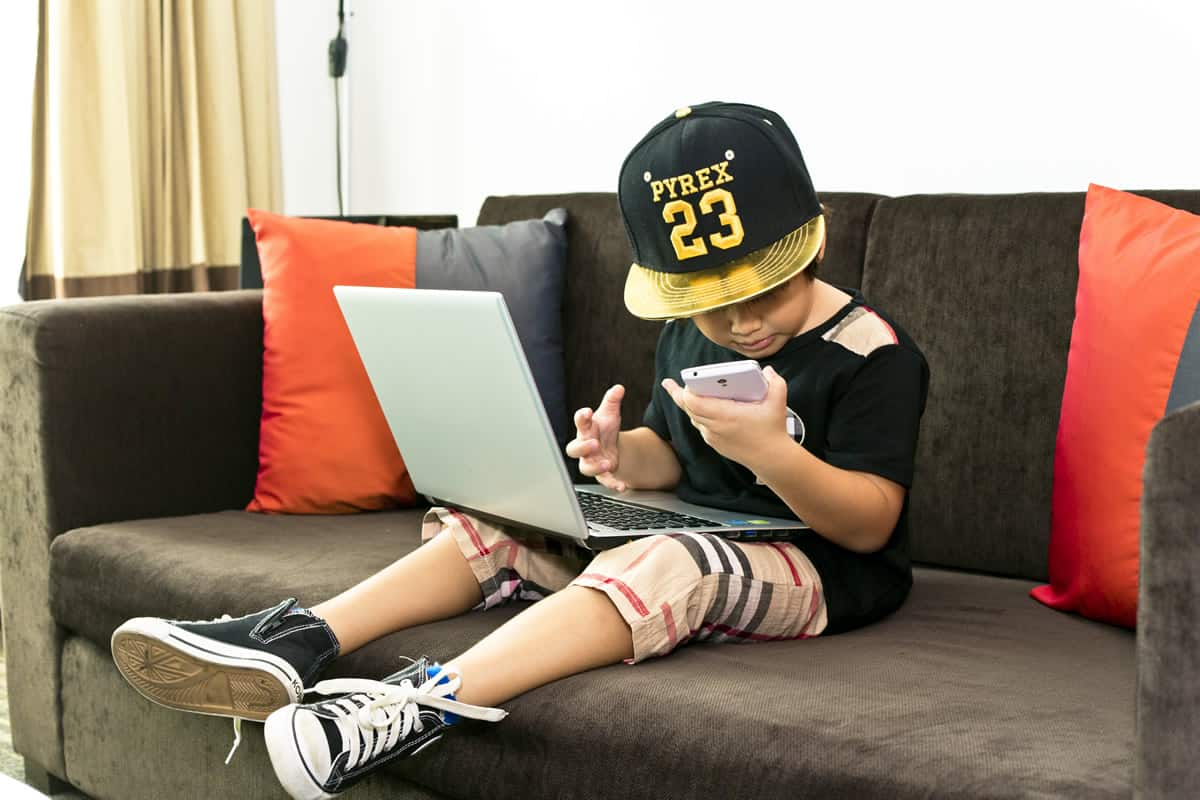 A photo of a child sitting with a laptop in his lap and a smartphone in his hand.