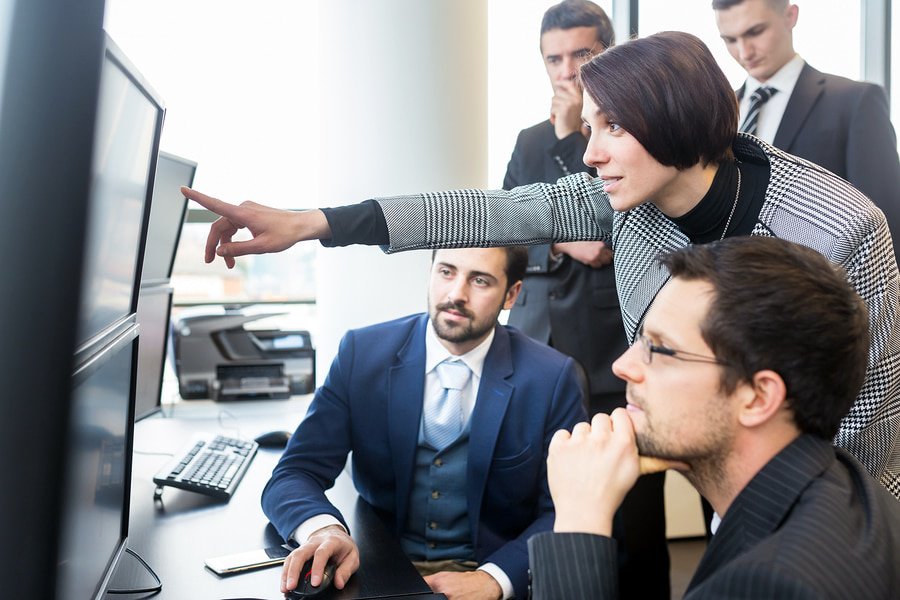  A group of people, including a woman in a suit, are gathered around a computer monitor discussing a product development plan.