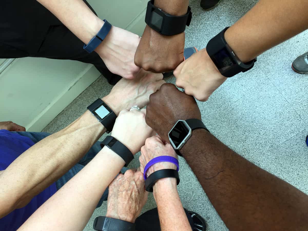 A photo of people putting their fists together in a circle, showing off their various Fitbit wearables.